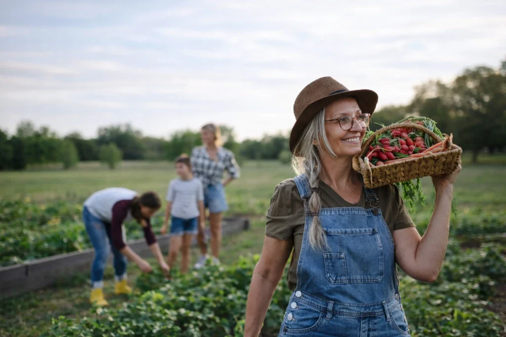 An older woman smiling while carrying a basket of freshly harvested vegetables in a community garden, with volunteers working together in the background.
