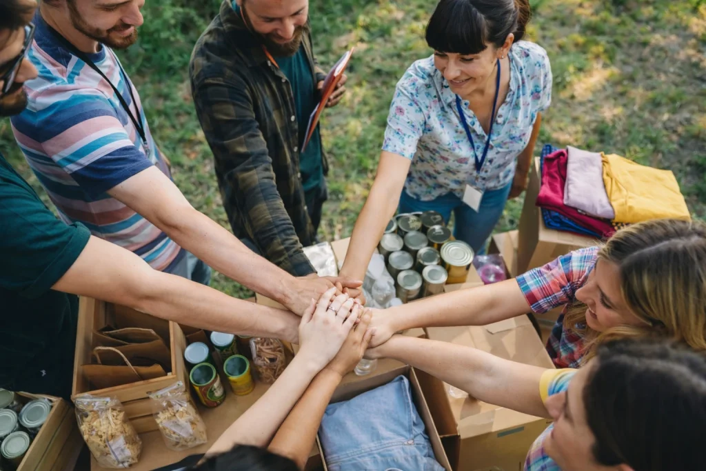 A group of volunteers standing in a circle with their hands stacked together over boxes of donated items outdoors.